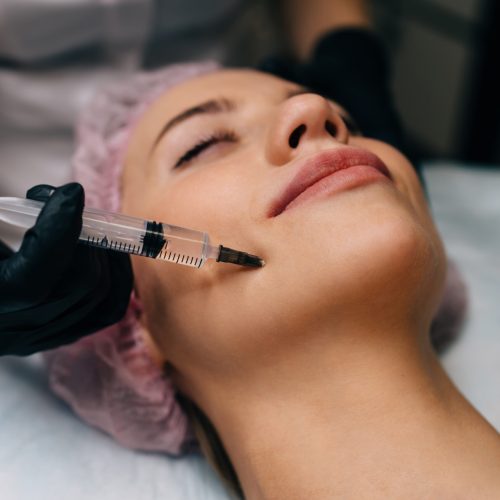 Cropped image of beautician doing botox injection with syringe to girl in beauty salon. Close up of young smiling caucasian woman client with closed eyes wearing medical cap. Beauty concept